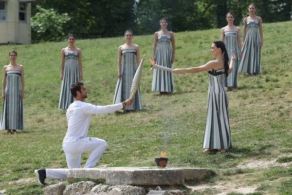 扮演最高女祭司的演員瑪麗·米娜（前右）點(diǎn)燃第一棒火炬手——希臘運(yùn)動(dòng)員、東京奧運(yùn)會(huì)男子賽艇項(xiàng)目金牌得主斯特凡諾斯·恩圖斯科斯手中的火炬。新華社記者 趙丁喆 攝